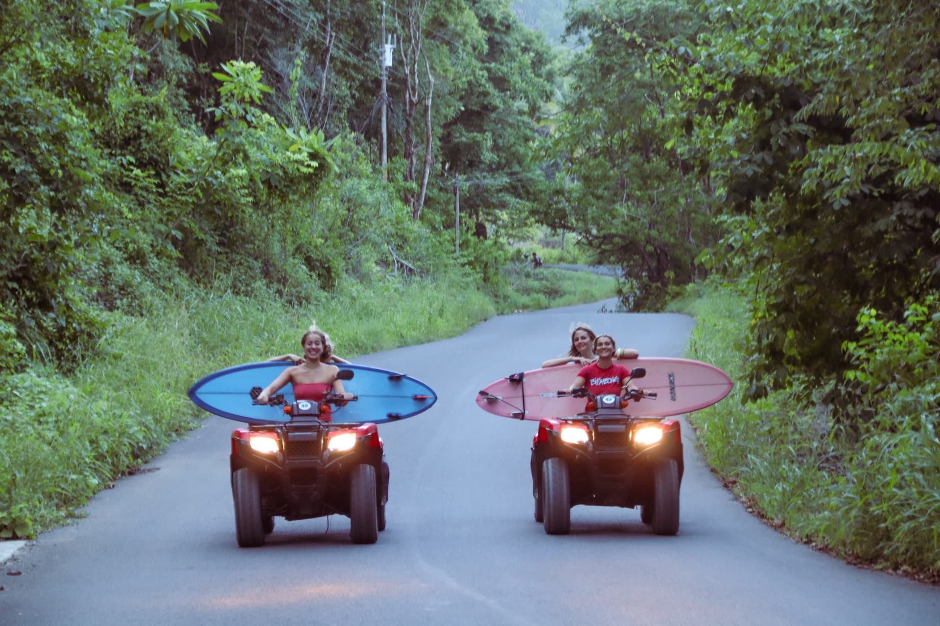 Two friends riding ATVs with surfboards down the road