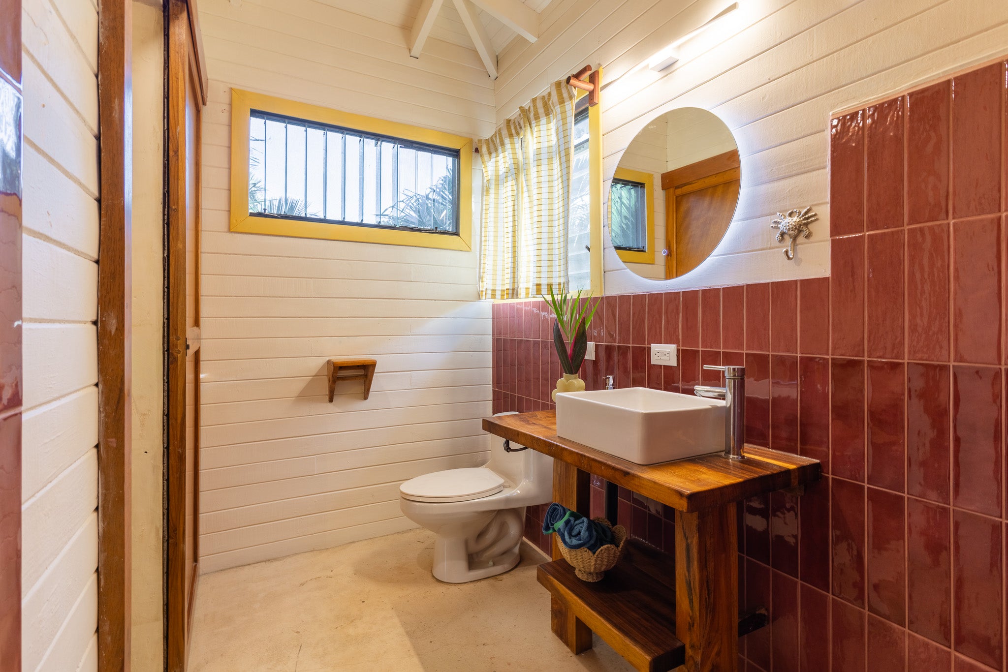 Studio Banano — Bathroom with red tiles, round lit mirror and wooden vanity