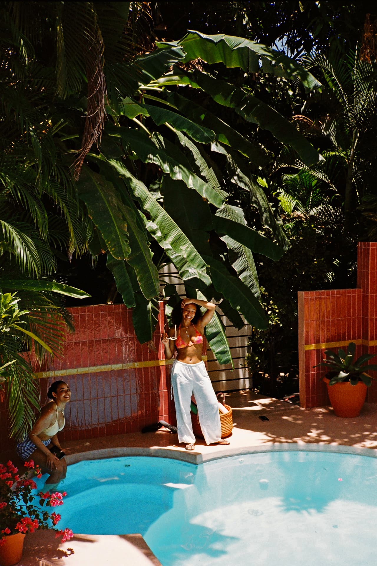 Two friends laughing poolside beneath the banana leaves